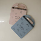 Two spiral-bound notebooks with pen holders, one pink and one blue, on a light beige background.
