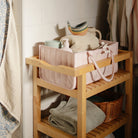 A wooden bathroom shelving unit holding a pink fabric storage caddy with bath products, topped with a set of stacked toy whales, with rolled towels stored on the lower shelf and hanging towels in the background.