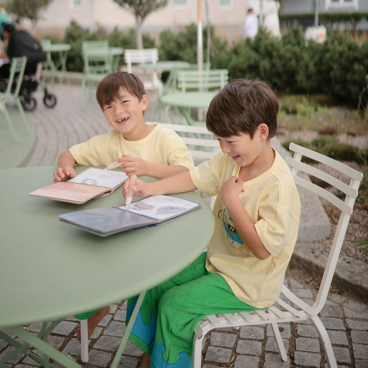 Two children sitting at a table outdoors, engaged with electronic devices.