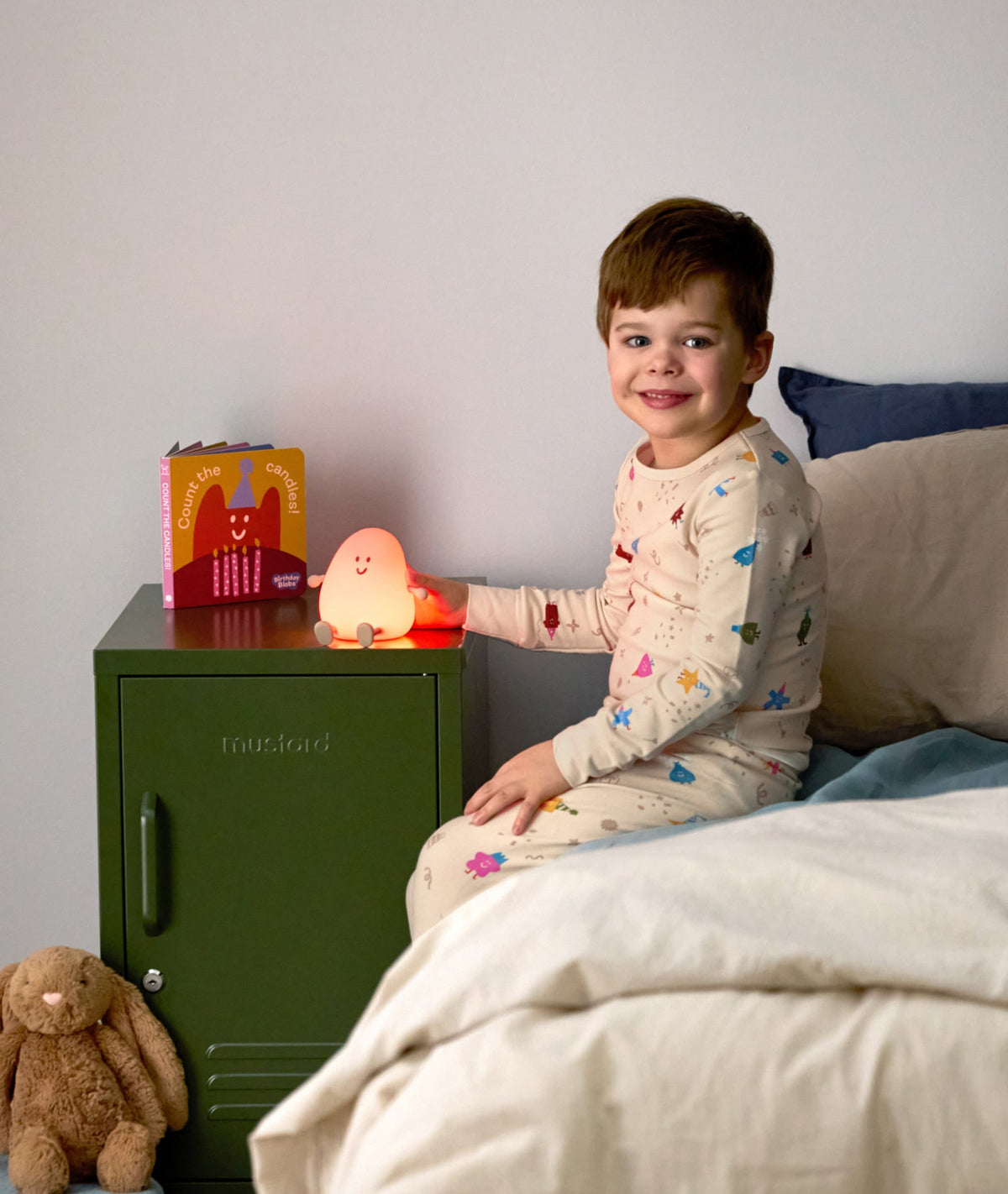 Child in pajamas sitting on a bed next to a nightstand with a night light and book