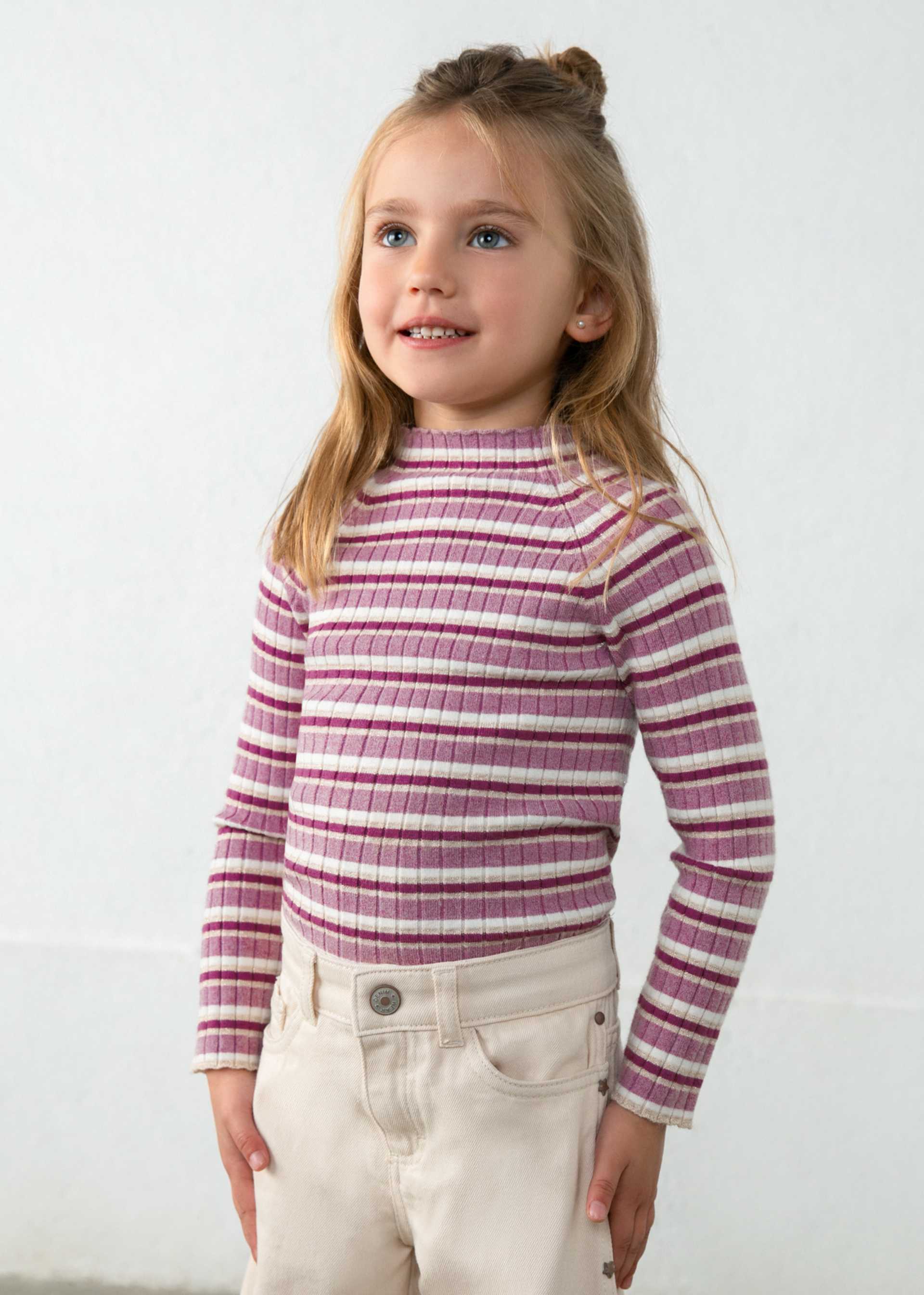 A young girl with blonde hair in a half-up bun wearing a ribbed mock neck turtleneck sweater with horizontal magenta and cream stripes, paired with cream pants, standing against a light gray wall.