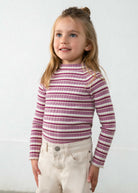 A young girl with blonde hair in a half-up bun wearing a ribbed mock neck turtleneck sweater with horizontal magenta and cream stripes, paired with cream pants, standing against a light gray wall.