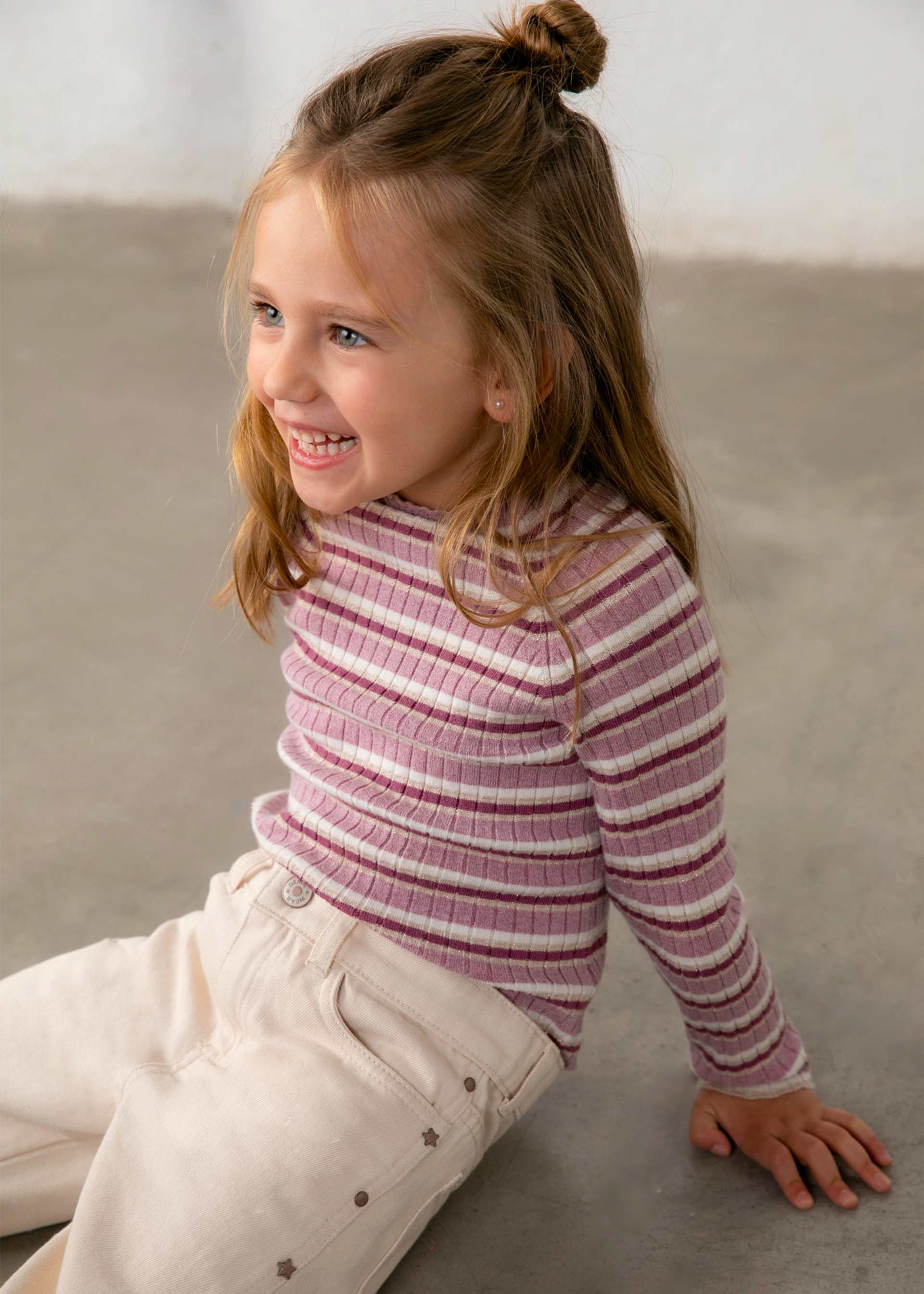 A young girl with a brown bun sits on a gray concrete floor wearing a pink and white striped ribbed turtleneck sweater and cream pants.