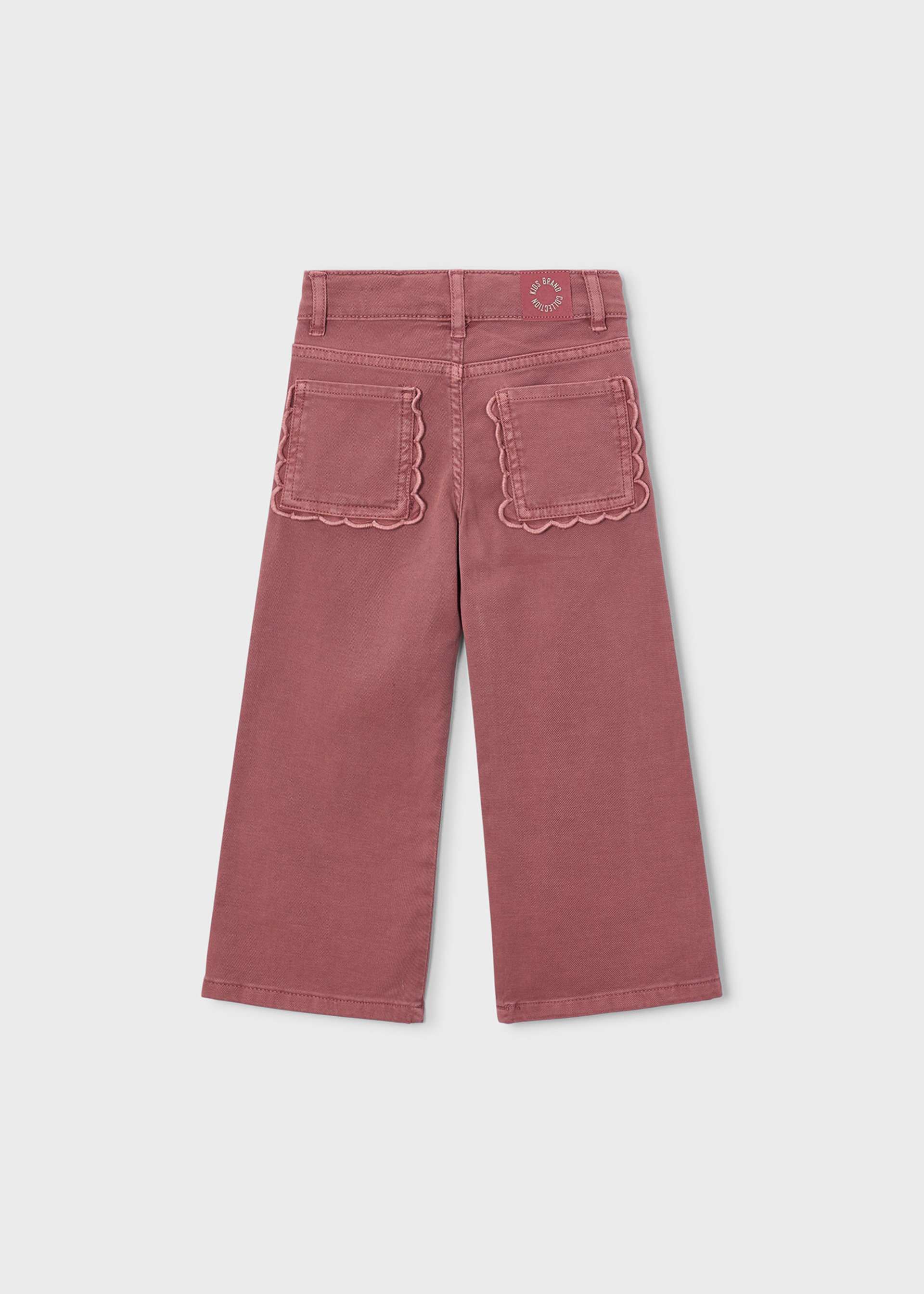 A pair of dusty rose cropped pants shown from the back, featuring scalloped edges on the back pockets and a small circular logo patch near the waistband, set against a light gray background.