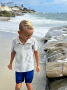 Child in a striped shirt and blue shorts standing on a beach with ocean waves in the background.