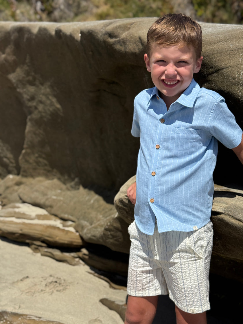 Young boy in a blue shirt and white shorts standing on a rocky surface.