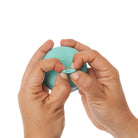 A close-up of a person's hands holding a small, round, mint green device and pressing a small release button on top, set against a solid white background.