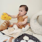 Baby in a mustard yellow romper sitting on a bed with toys and blankets.