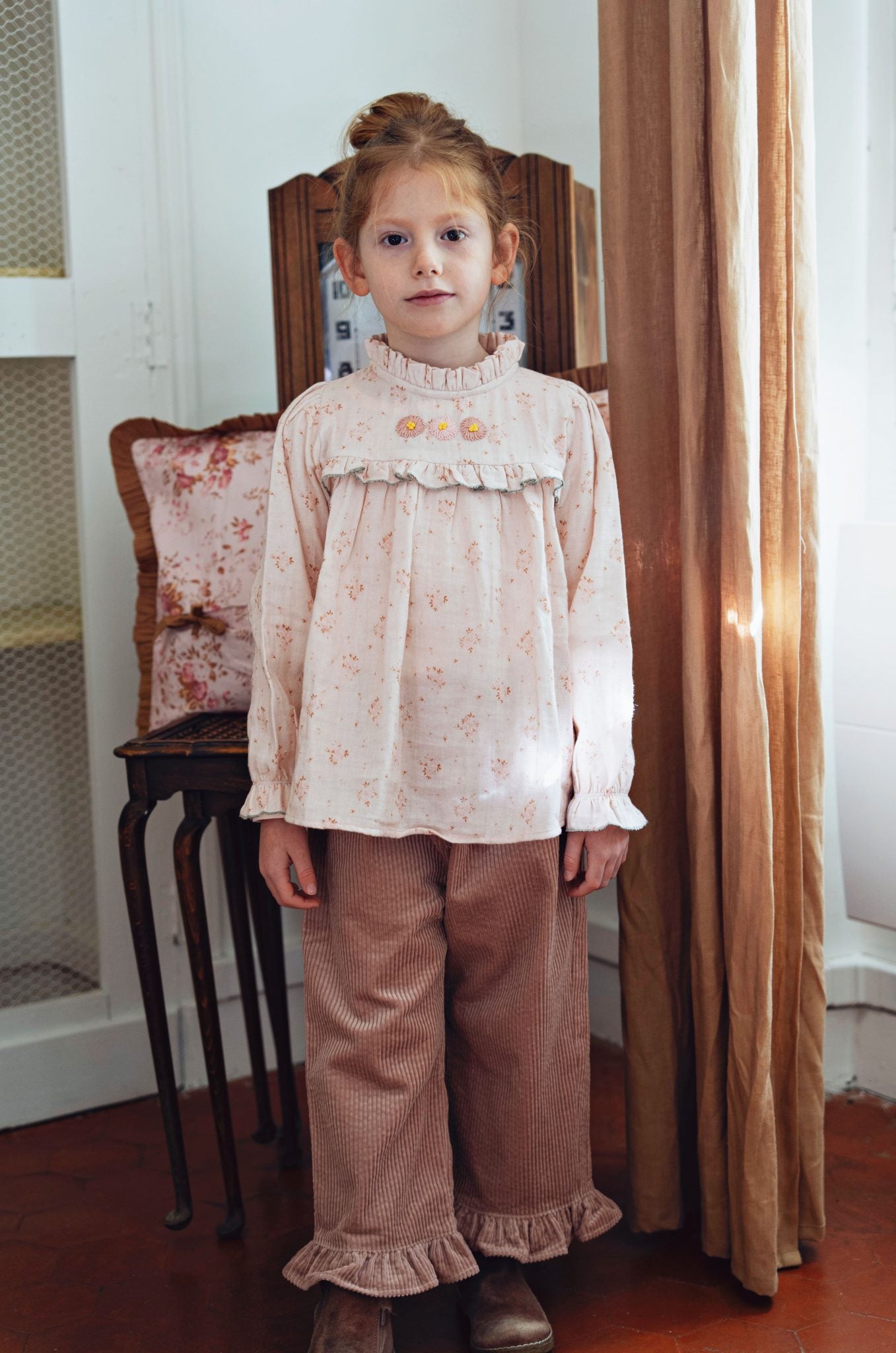 A young girl stands indoors wearing a light pink long-sleeve blouse with a ruffled yoke and small floral print, paired with brown corduroy pants with ruffled hems. She is posed in front of a window with warm sunlight, a wooden chair, and a curtain.