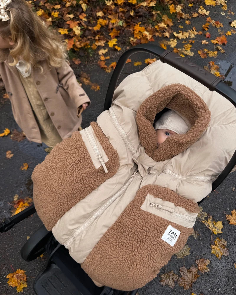 A young child with a plaid hair bow reaches down toward an infant car seat where a baby is sleeping inside a Benji Applique Car Seat Cocoon by 7AM Enfant in oat with brown sherpa fleece trim, outdoors near a paved path scattered with autumn leaves.