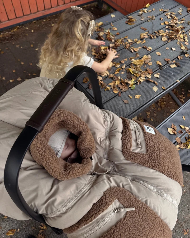 A young child with long blonde hair reaches down toward an infant car seat where a baby is sleeping inside a Benji Applique Car Seat Cocoon by 7AM Enfant in oat with brown sherpa fleece trim, outdoors near a wooden bench scattered with autumn leaves.