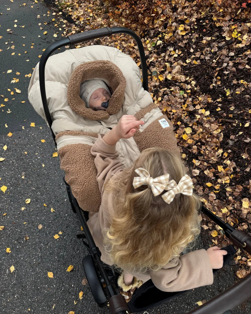 A young child with a plaid hair bow reaches down toward an infant car seat where a baby is sleeping inside a Benji Applique Car Seat Cocoon by 7AM Enfant in oat with brown sherpa fleece trim, outdoors near a paved path scattered with autumn leaves.
