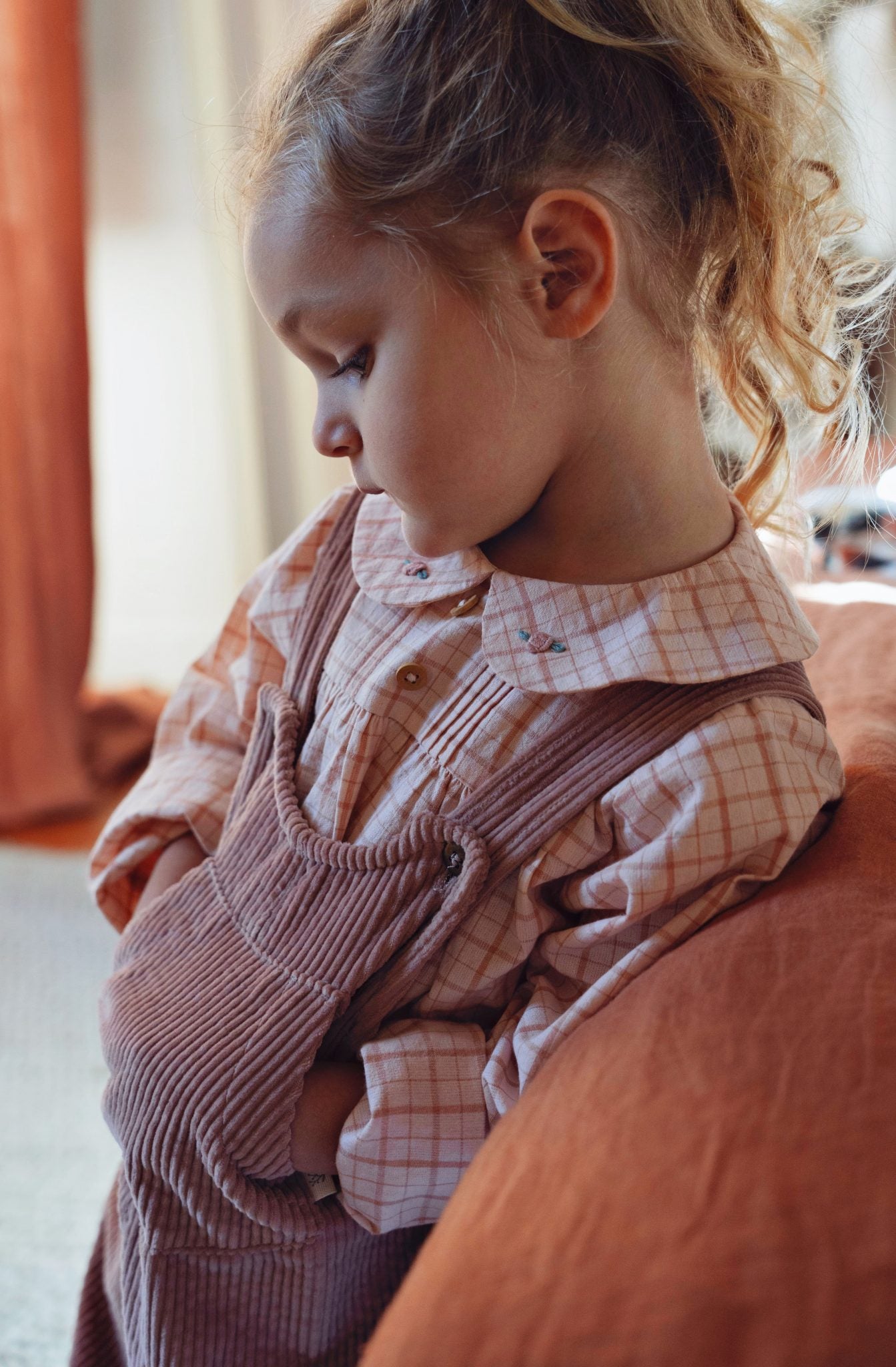 A young child wearing a pink and white checkered long-sleeve shirt under a dusty rose corduroy overall, sitting on a rust-colored couch in a softly lit room.