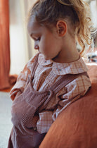A young child wearing a pink and white checkered long-sleeve shirt under a dusty rose corduroy overall, sitting on a rust-colored couch in a softly lit room.