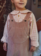 A child wearing a pink and white checkered long-sleeve shirt under a dusty rose corduroy overall dress with a Peter Pan collar and gold buttons.