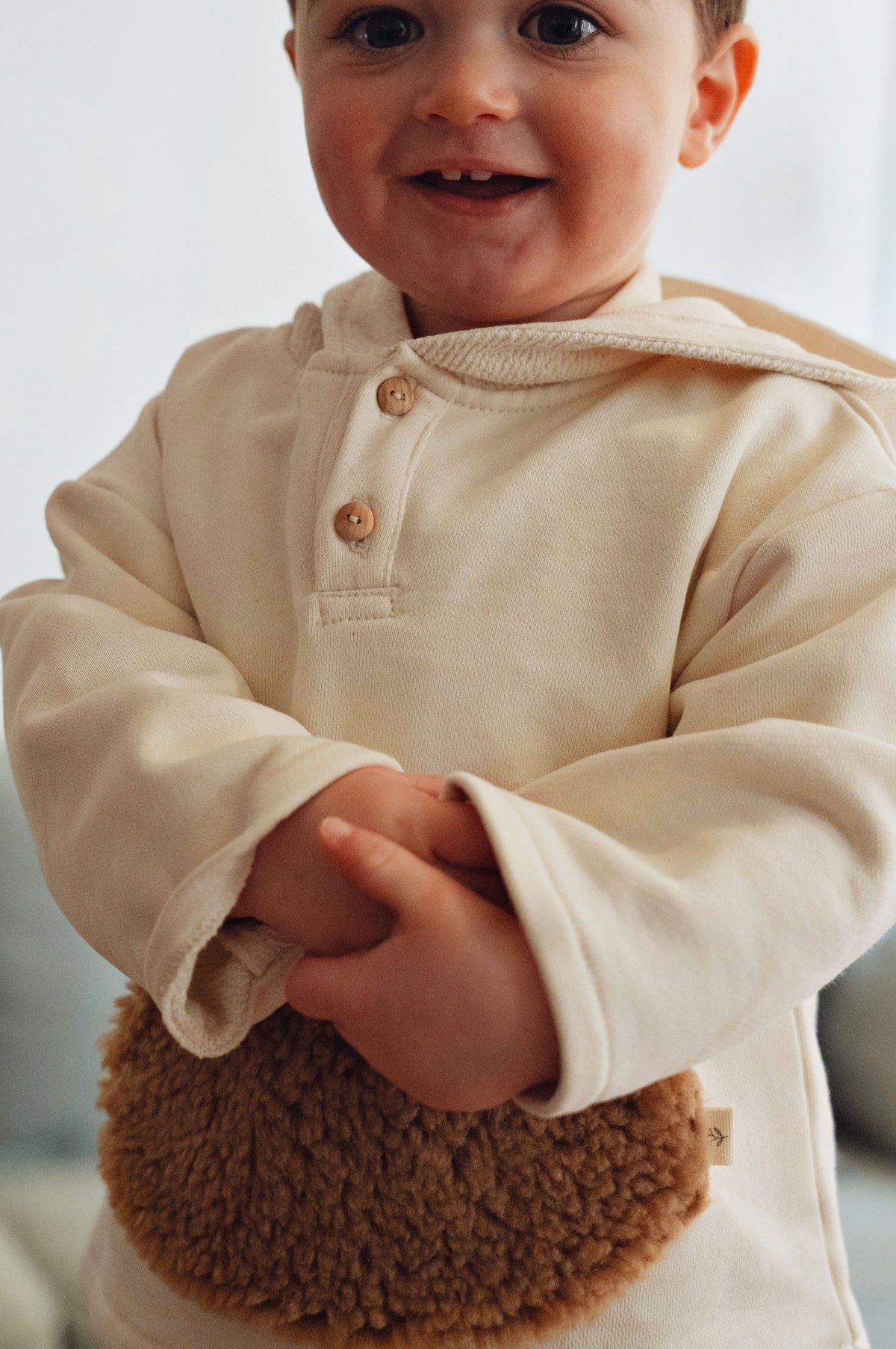 A close-up of a smiling baby wearing a cream-colored long-sleeve Henley top with wooden buttons, holding a brown textured plush pouch.