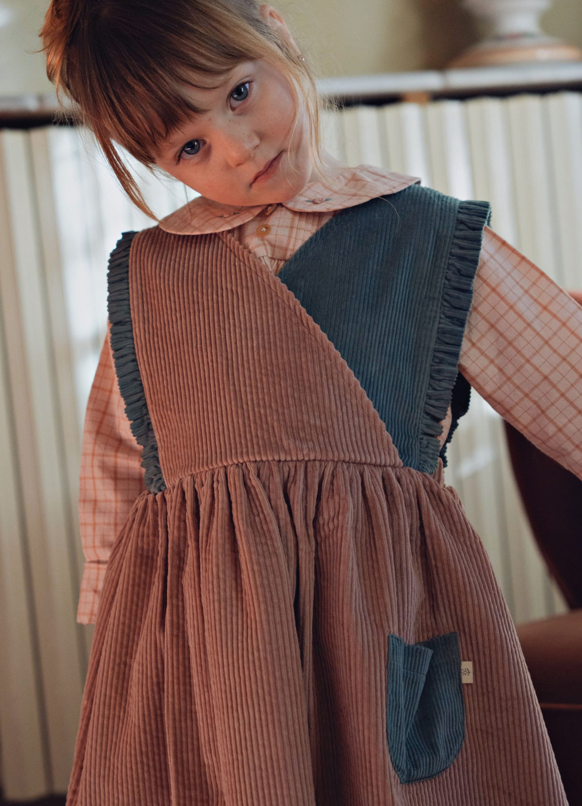 A young child wearing a brown corduroy pinafore dress over a peach plaid collared shirt, photographed indoors against a softly blurred background.