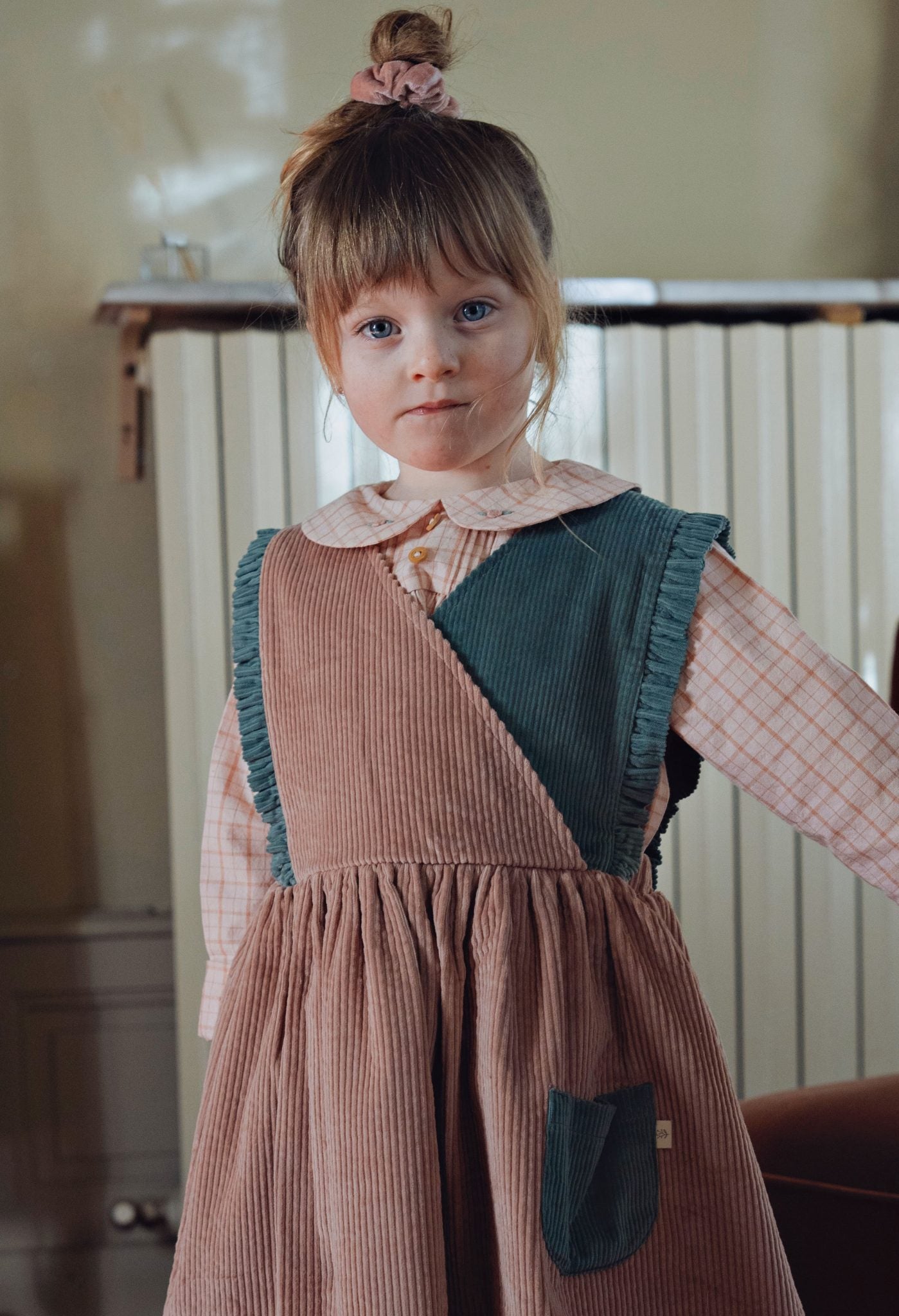A young girl with a messy bun and blue eyes wearing a brown corduroy pinafore dress with a dark green panel and a pink gingham shirt underneath, standing in an indoor setting.
