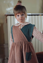 A young girl with a messy bun and blue eyes wearing a brown corduroy pinafore dress with a dark green panel and a pink gingham shirt underneath, standing in an indoor setting.