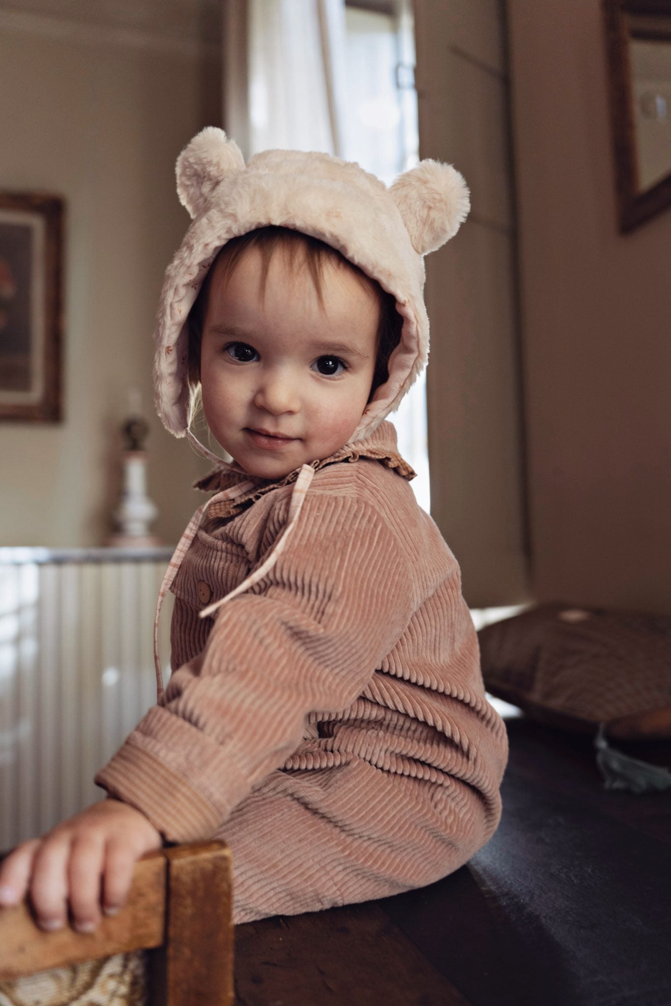 A baby wearing a light beige plush bear-ear bonnet and a dusty rose corduroy romper, sitting on a wooden chair in a softly lit indoor room.