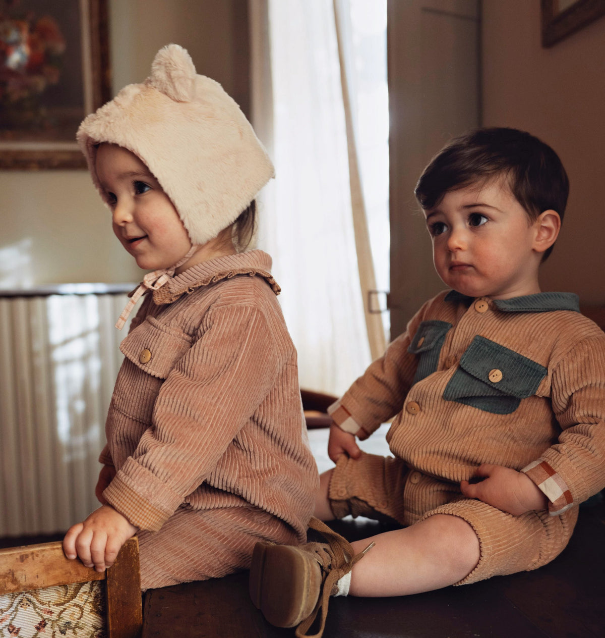 Two toddlers sitting indoors wearing matching corduroy outfits. The child on the left wears a beige fuzzy beanie with bunny ears and a tan corduroy jumpsuit with a pink bow.
