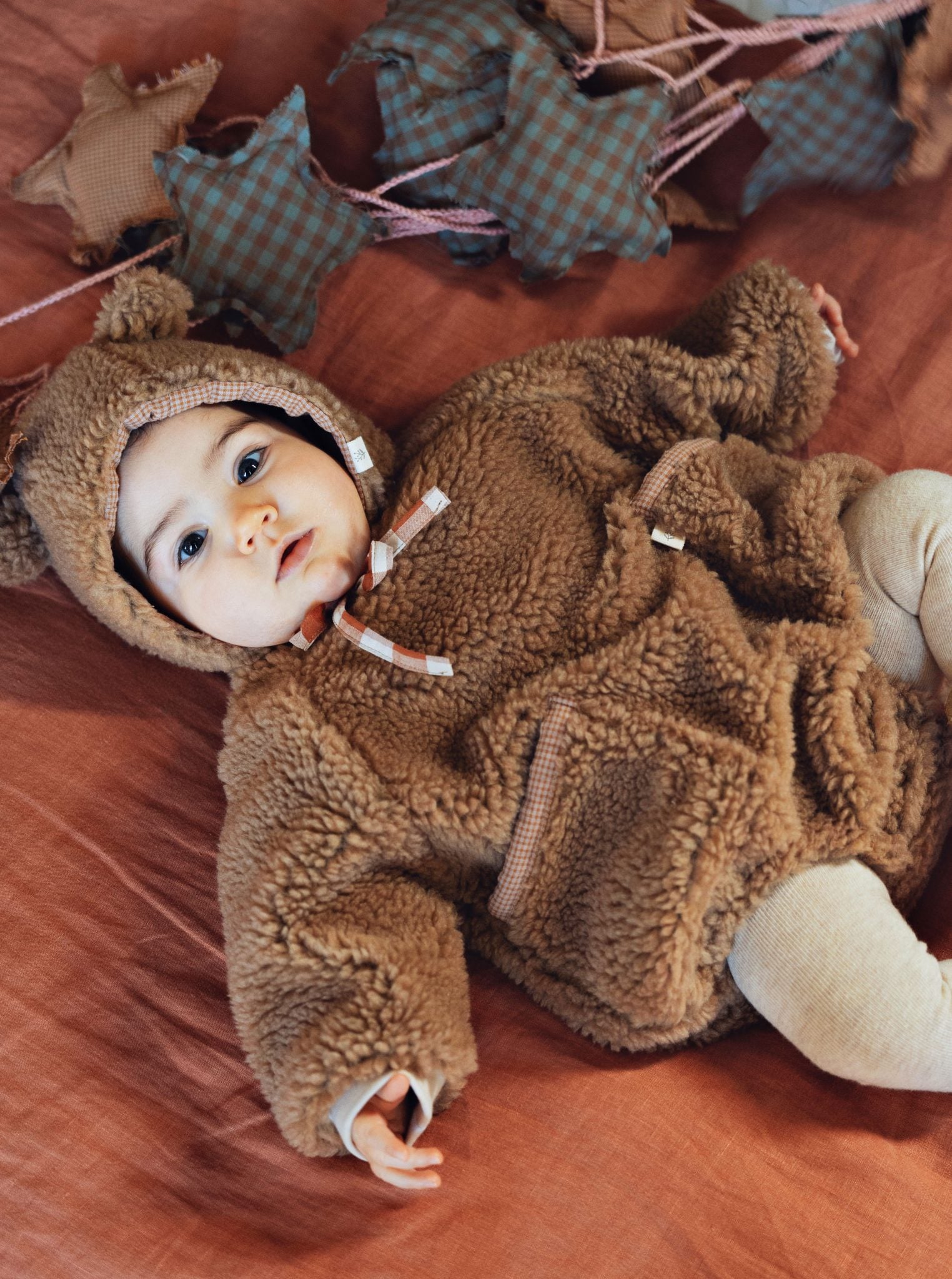 A baby lying on a rust-colored fabric surface wearing a brown teddy fleece hooded outfit with ear details and drawstrings. A matching brown fleece hood is also shown nearby, along with soft star-shaped pillows and a plush toy in the background.