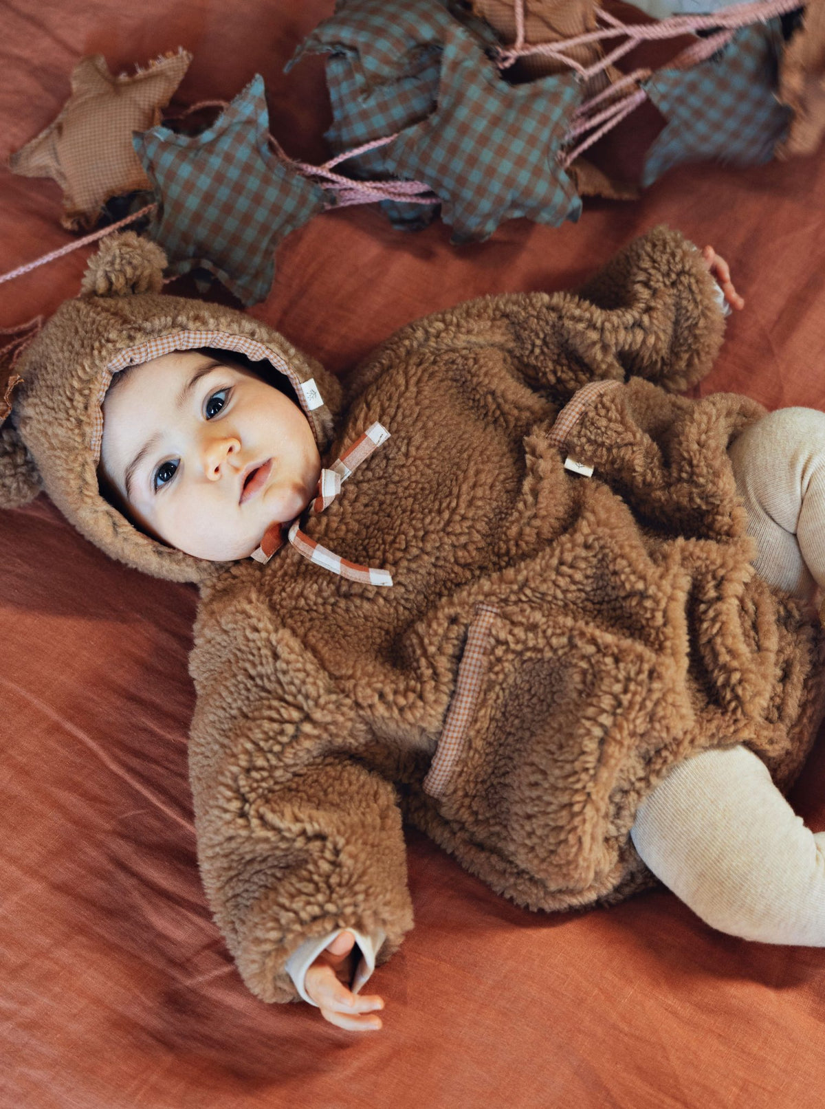 A baby lying on a rust-colored fabric surface wearing a brown teddy fleece hooded outfit with ear details and drawstrings. A matching brown fleece hood is also shown nearby, along with soft star-shaped pillows and a plush toy in the background.