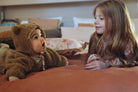 A baby and a girl  lying on a rust-colored fabric surface wearing a brown teddy fleece hooded outfit with ear details and drawstrings. A matching brown fleece hood is also shown nearby.background.