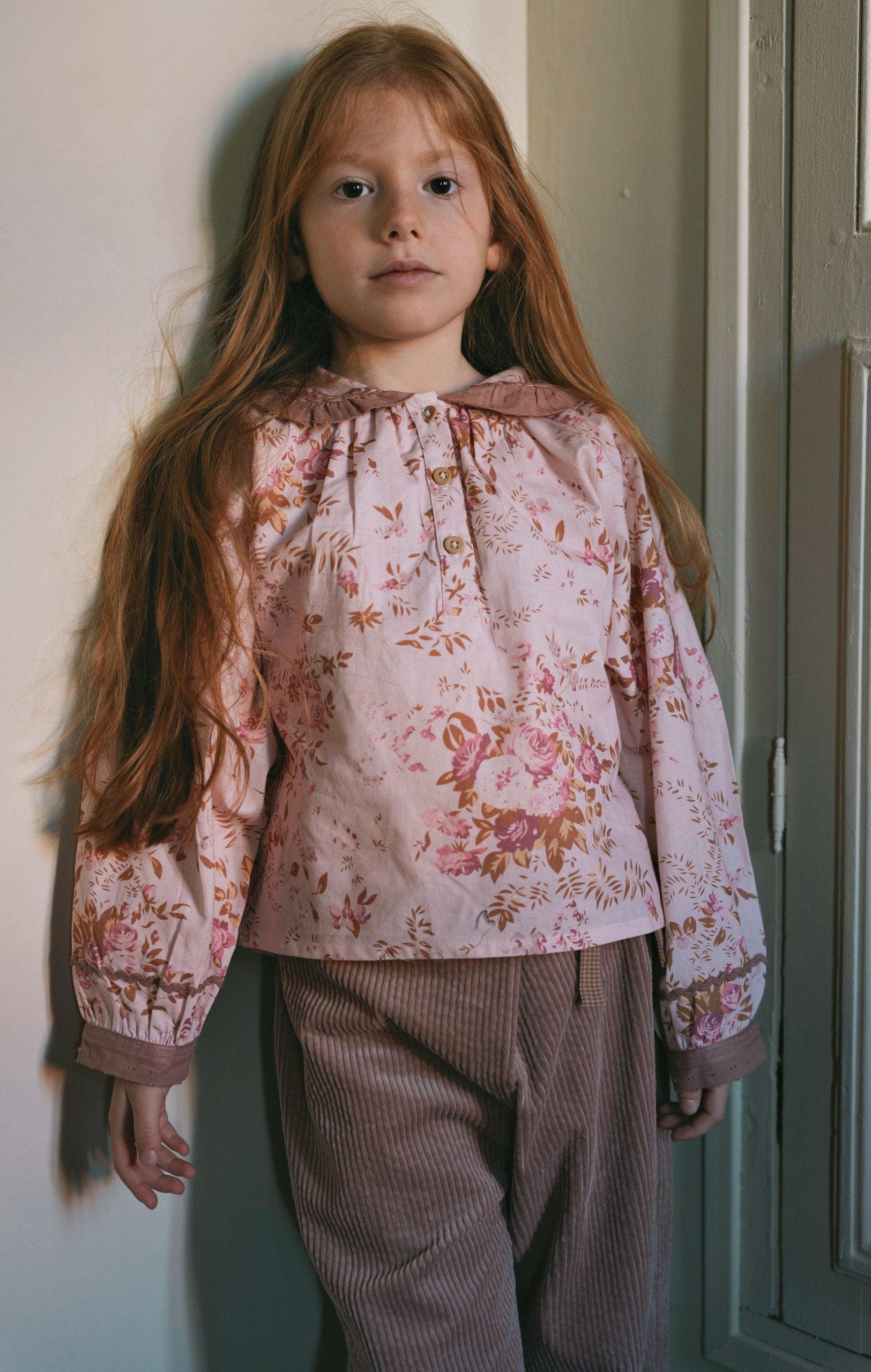 A child wearing a dusty rose eyelet blouse with a cream lace collar and matching ruffled cuffs, paired with dark green corduroy pants.