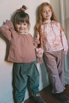 Two young girls standing. The girl in right wears a light pink floral long-sleeve blouse with ruffled trim and loose gray trousers, while the girl in the right wears a dusty pink blouse and dark green trousers.