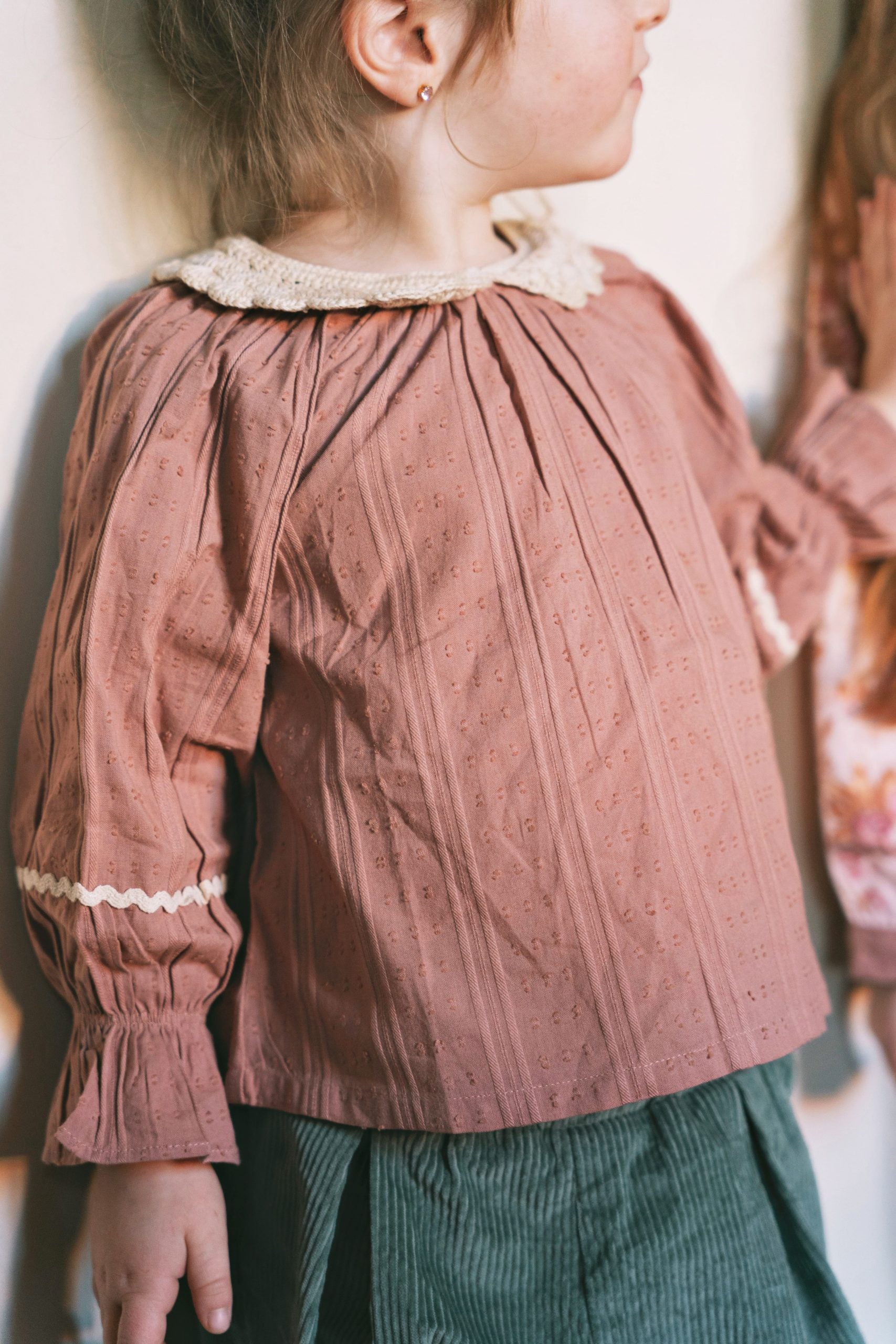A child wearing a dusty rose eyelet blouse with a cream lace collar and matching ruffled cuffs, paired with dark green corduroy pants.