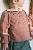 A child wearing a dusty rose eyelet blouse with a cream lace collar and matching ruffled cuffs, paired with dark green corduroy pants.
