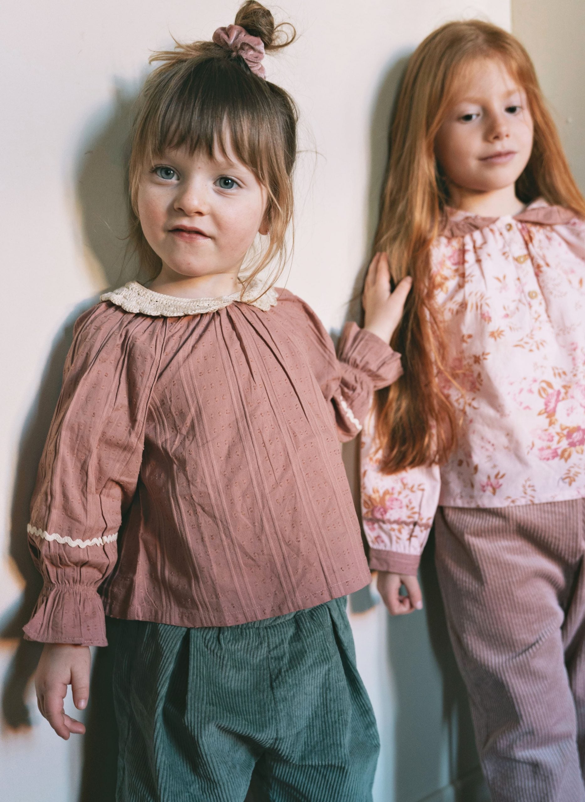 Two young girls wearing vintage-style clothing, standing against a plain wall. The girl in the foreground wears a dusty rose long-sleeve top with a cream lace collar and dark green corduroy pants, while the girl behind her wears a light pink floral top and matching corduroy pants.