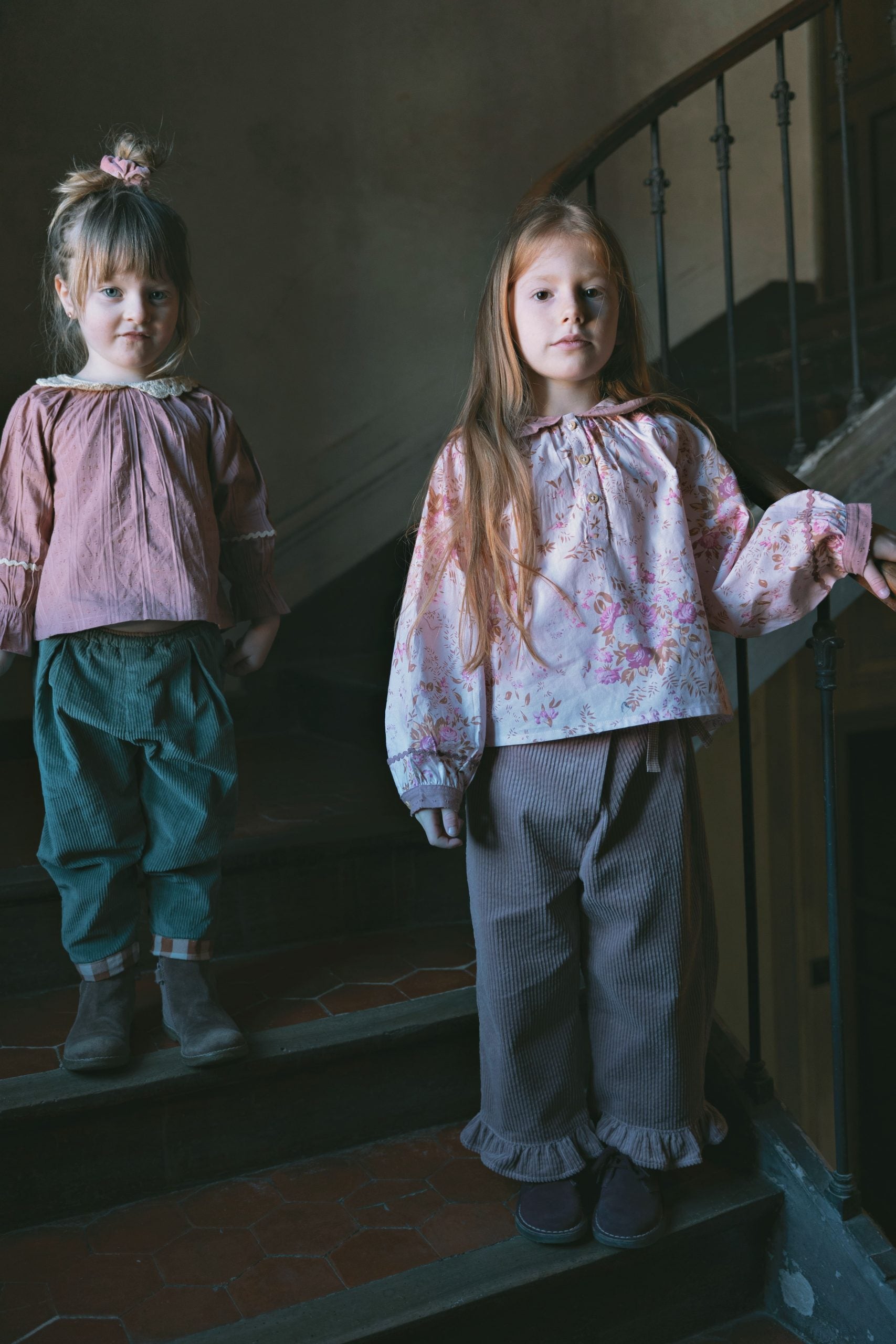 Two young girls standing on an old stone staircase indoors. The girl in front wears a light pink floral long-sleeve blouse with ruffled trim and loose gray trousers, while the girl behind wears a dusty pink blouse and dark green trousers.