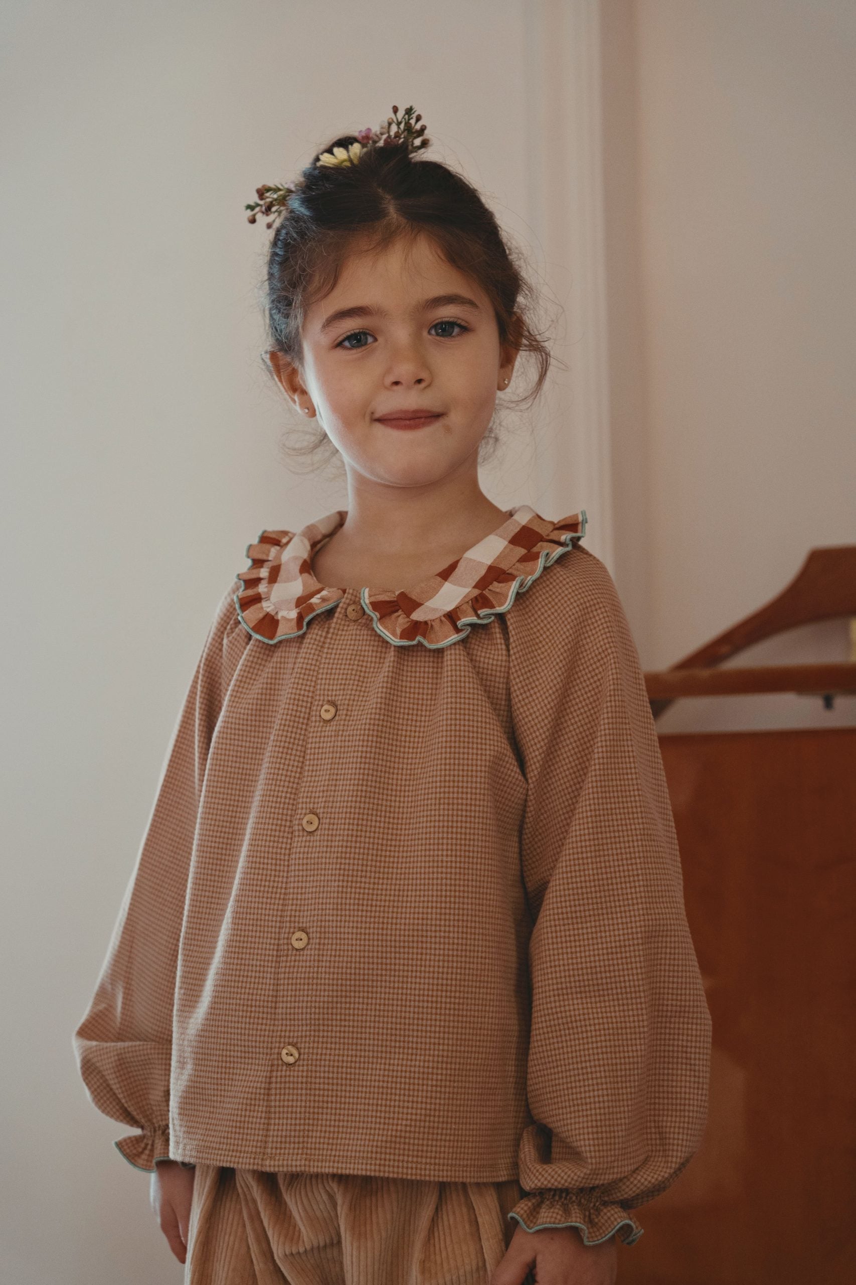 A young girl with her hair in a small floral crown stands indoors wearing a brown gingham long-sleeve button-up shirt with a ruffled collar in brown, beige, and white, paired with matching brown gingham trousers.