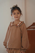 A young girl with her hair in a small floral crown stands indoors wearing a brown gingham long-sleeve button-up shirt with a ruffled collar in brown, beige, and white, paired with matching brown gingham trousers.