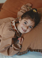 A young girl with dark hair and a small flower hair accessory lies on a brown checkered pillow, wearing a matching brown checkered long-sleeve top with a ruffled collar.