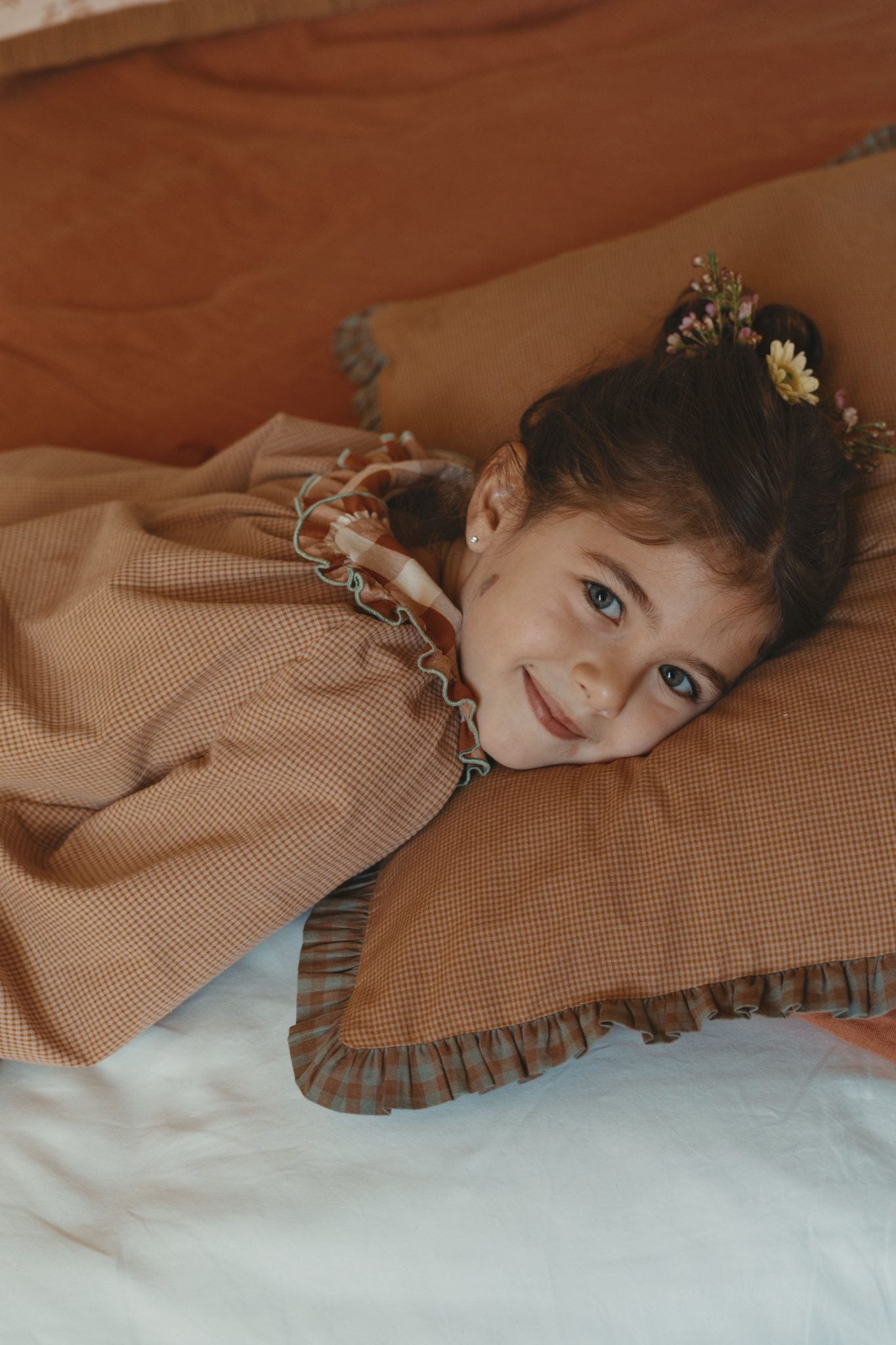 A young girl with a floral hair accessory lies on a bed, smiling at the camera. She is wearing a textured brown long-sleeve top with a ruffled collar, resting on a matching brown pillow against a warm brown background.