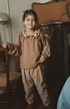 A young girl wearing a vintage-style brown corduroy pajama set with a patterned ruffled collar, standing in a cozy, warm-toned room.
