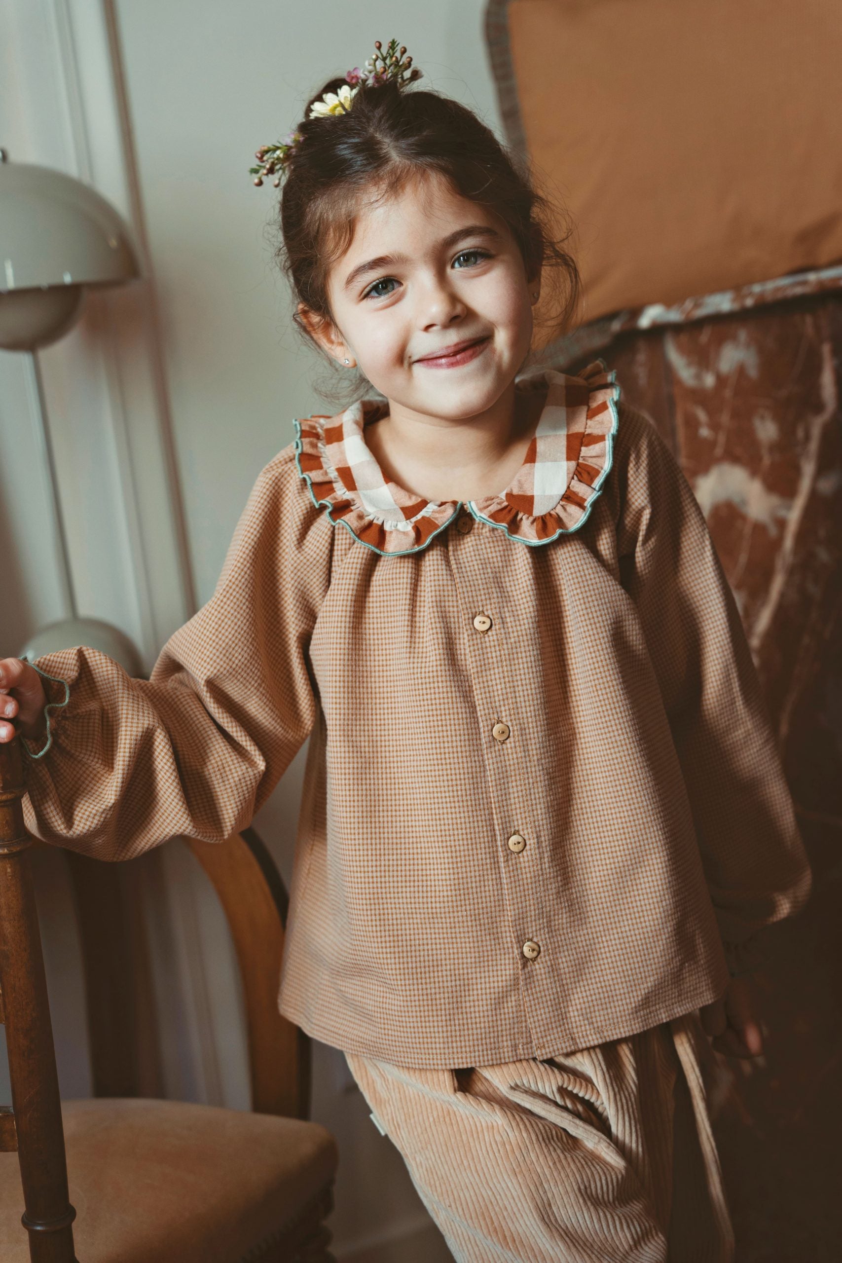 A young girl with her hair in a small floral crown stands indoors wearing a brown gingham long-sleeve button-up shirt with a ruffled collar in brown, beige
