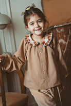 A young girl with her hair in a small floral crown stands indoors wearing a brown gingham long-sleeve button-up shirt with a ruffled collar in brown, beige