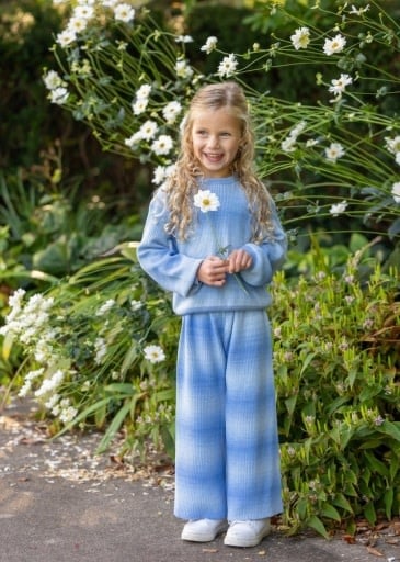 A young girl with blonde hair stands in a garden wearing a matching light blue sweatshirt and wide-leg pants set with a subtle vertical gradient. She is holding a small white daisy flower.