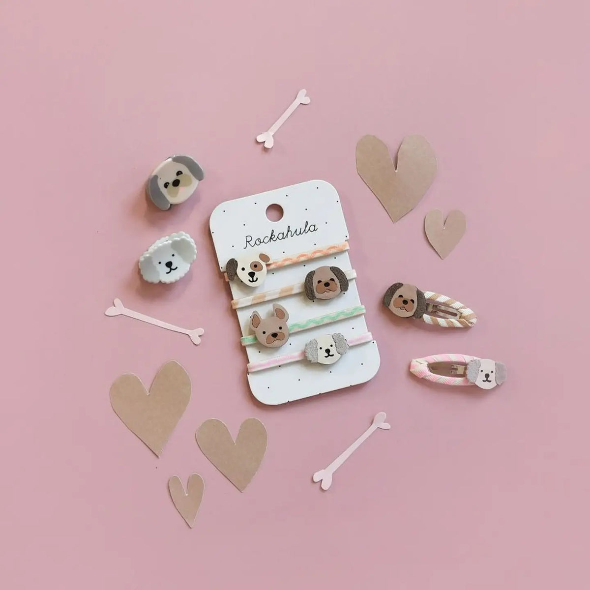 A set of dog-themed hair accessories displayed on a pink background with decorative paper hearts and bones. The set includes a white display card labeled 'Rockahula' with four elastic hair ties featuring dog faces, plus three matching hair clips.