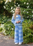 A young girl with blonde hair stands in a garden wearing a matching light blue sweatshirt and wide-leg pants set with a subtle vertical gradient. She is holding a small white daisy flower.