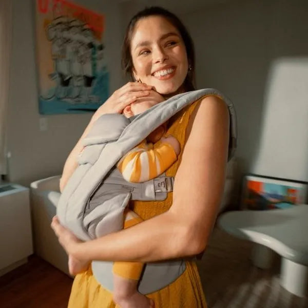 Woman holding a baby in a carrier in a room with a lamp and artwork on the wall.