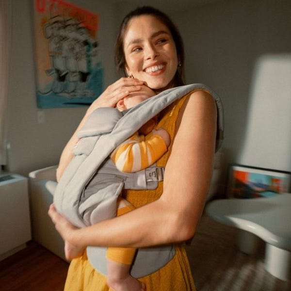 Woman smiling while holding a baby in a sling indoors.