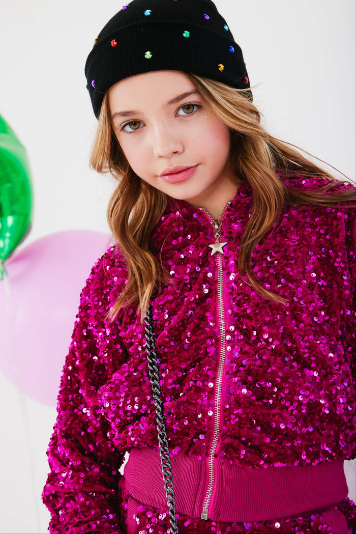 A young girl wearing a sparkling magenta sequined zip-up jacket, a matching pink top, a black beanie with colorful rhinestones, and holding a silver chain bag, set against a white background with balloons.