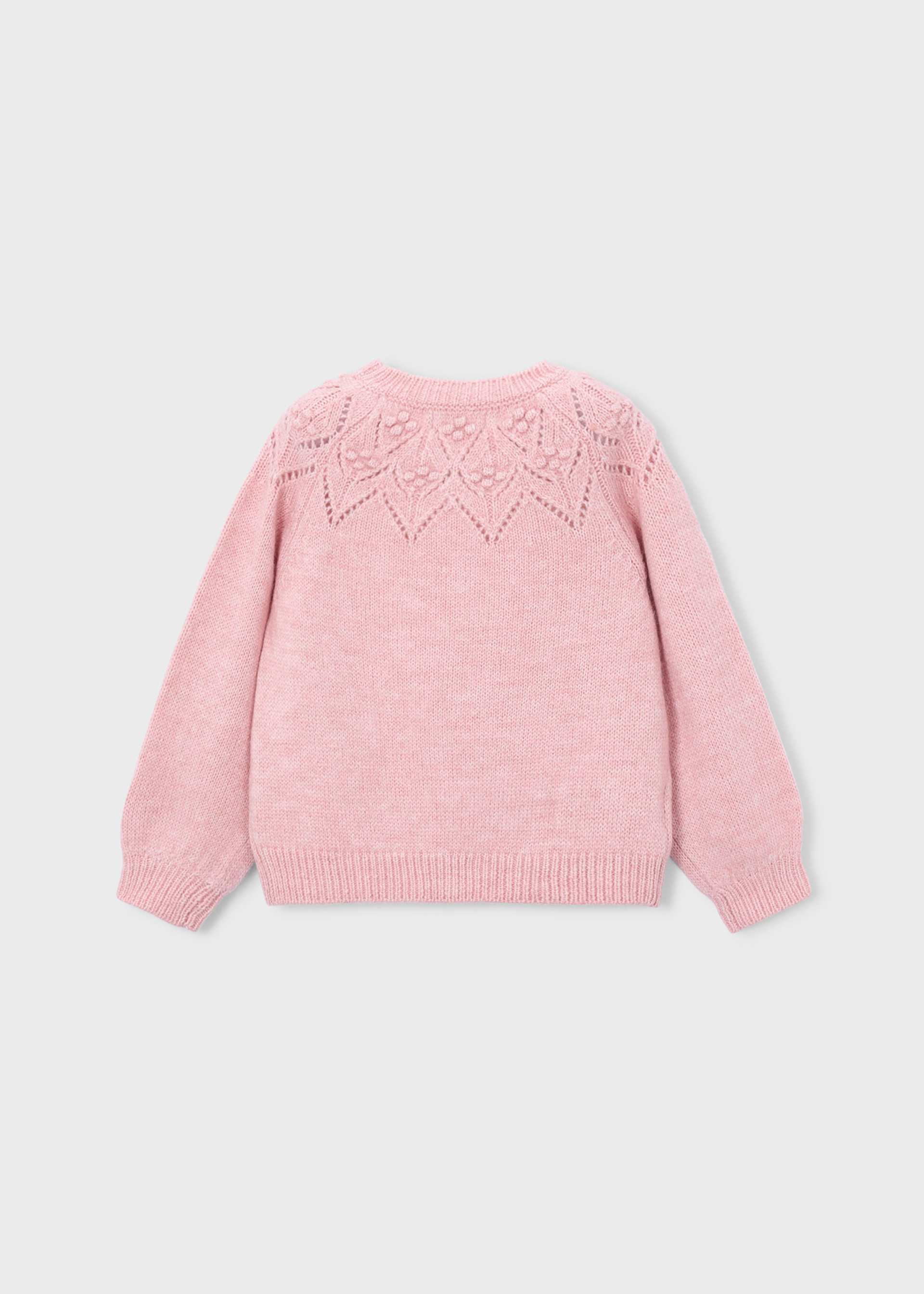 A light pink knit sweater shown from the back, featuring a delicate geometric lace-knit pattern across the upper back, set against a solid light gray background.