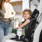 Child in a car seat with a woman standing nearby, inside a vehicle.