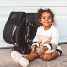 Child sitting next to a car seat with a visible brand logo on a neutral background